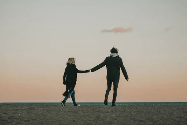 Mann und Frau gehen am Strand Sand bei Sonnenuntergang, Händchen haltend, mit dem Meer und einem blauen Himmel im Hintergrund. — Stockfoto