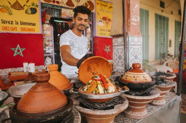 Rissani, Fas - 18 Eylül 2019: Genç Faslı adam sokakta sebzelerle Taine pişiriyor ve Rissani, Fas 'ta yemek satması için turistlere gösteriyor.