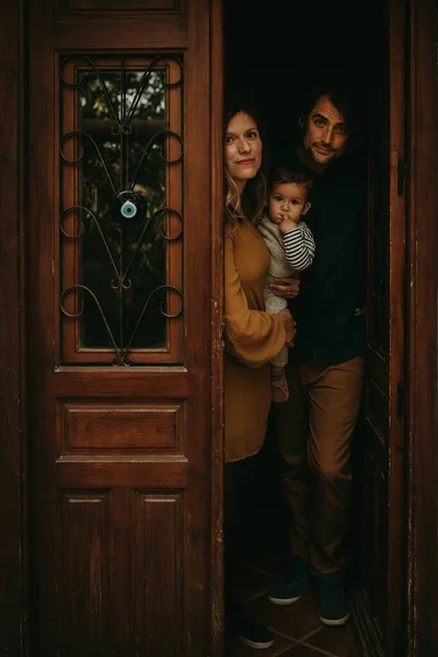 Family portrait of young man and woman holding their baby son at the entrance door of their home, with beautiful low light.