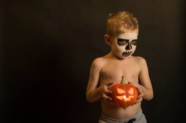 Cadılar Bayramı makyajında ellerinde balkabağı olan küçük çocuk sarı bir arka planda.