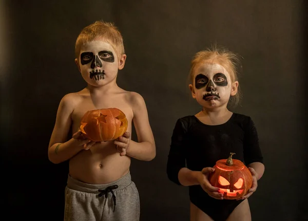 Bambini un ragazzo e una ragazza con le zucche nelle loro mani nel trucco Halloween guardare la fotocamera su uno sfondo scuro Fotografia Stock