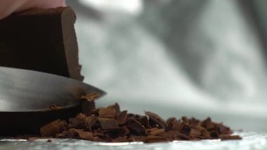 Çikolata parçacıkları, arka planda gümüş bir arka plan videosundaki çikolata çubuğundan keskin bir bıçakla kesilmiş.