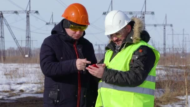 Werknemer en power engineer aan het praten op de achtergrond van een hoogspanningslijn, kijk naar een smartphone — Stockvideo