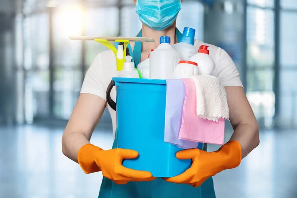 A cleaning lady in a mask holds a bucket of cleaning products on the background of windows.