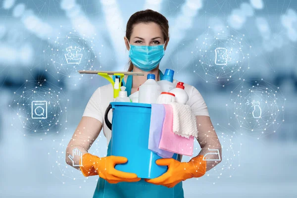The concepts of cleaning and disinfection services.A cleaning lady in a mask stands on a blurry background with cleaning products.