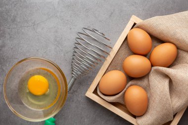 Tahta tepside tavuk yumurtası, yumurta çırpıcı ve siyah beton zeminde cam kase. Üst görünüm.