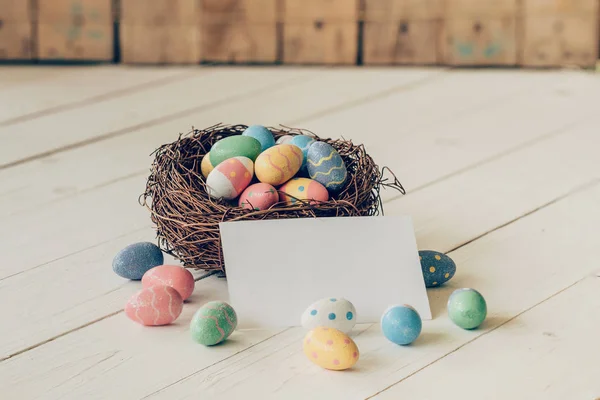 Красочные пасхальные яйца в гнезде и бумажная карточка на деревянном столе ба — стоковое фото