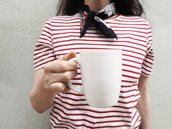Weiße Becher-Attrappe vorhanden. Nahaufnahme einer jungen Frau in gestreiftem T-Shirt und Schal, die eine Tasse Tee oder Kaffee in der Hand hält. Weibliche Aktienfotografie. Frontansicht. — Stockfoto