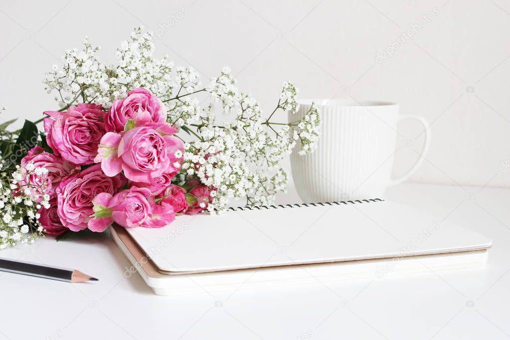 Wedding styled stock photo. Still life with pink roses, babys breath Gypsophila flowers, white cup, pencil and notebook. Floral composition. Image for blog or social media.