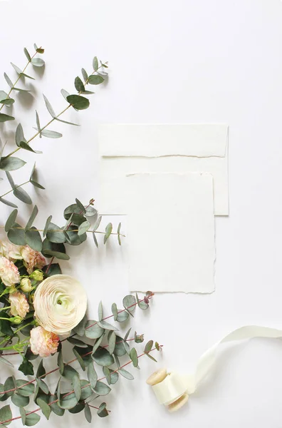 Matrimonio femminile, modelli di desktop di compleanno. Biglietto di auguri in carta bianca, busta. Rami di eucalipto, rose rosa e fiori di ranuncolo persiano. Sfondo tavolo bianco. Posa piatta, vista dall'alto . — Foto Stock