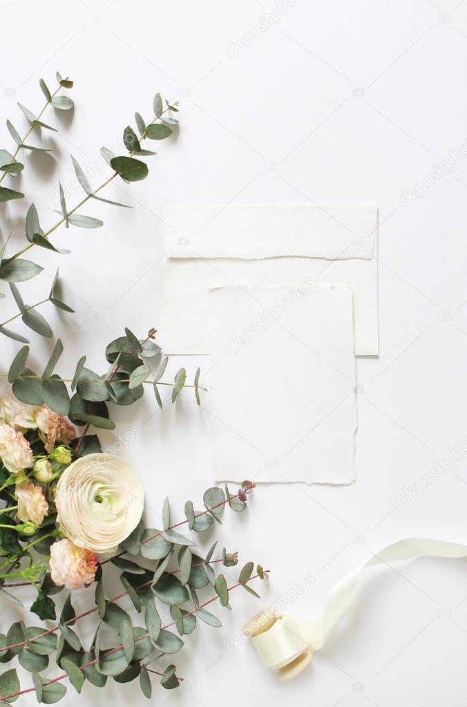 Feminine wedding, birthday desktop mock-ups. Blank craft paper greeting card, envelope. Eucalyptus branches, pink roses and Persian buttercup flowers. White table background. Flat lay, top view.