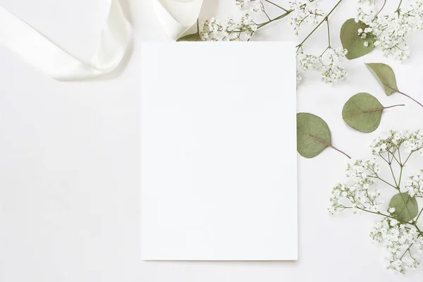 Στυλ φωτογραφία απόθεμα. Θηλυκή γάμου χαρτικά γραφείου mockup με κενό ευχετήρια κάρτα, babys ανάσα γυψοφίλης λουλούδια, φύλλα ξηρό πράσινο ευκάλυπτο, σατέν κορδέλα και λευκό φόντο. Κενός χώρος — Φωτογραφία Αρχείου