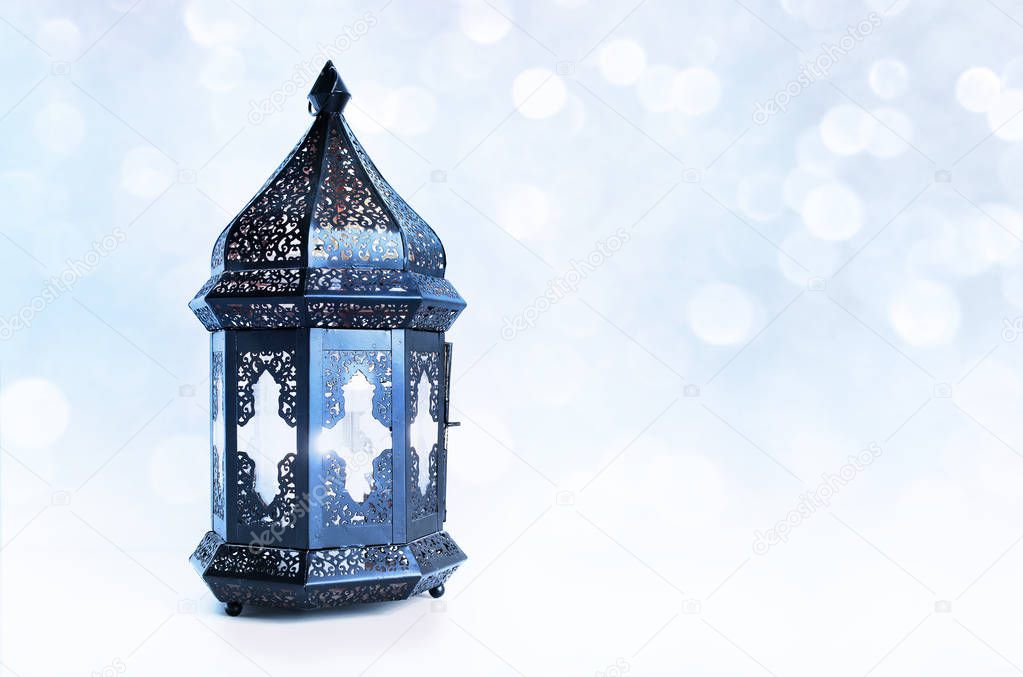 Ornamental dark Moroccan, Arabic lantern on the table. Burning candle, glittering bokeh lights stars. Greeting card for Muslim community holy month Ramadan Kareem. Festive blue blurred background.