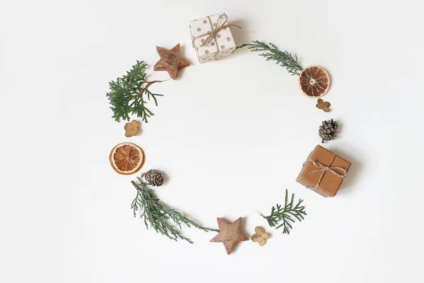 Christmas circle floral composition. Wreath of juniperus branches, pine cones, gift boxes, wooden stars and dry orange fruit on white background. Winter, advent design and decoration. Flat lay, top