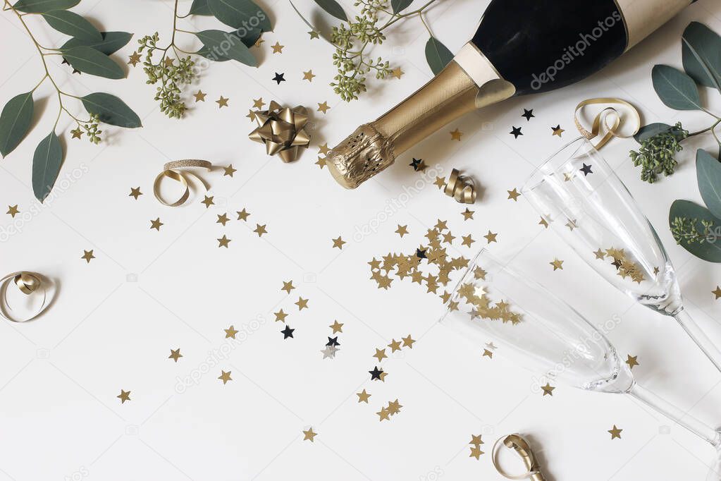Happy New Year composition. Champagne glasses nad wine bottle with golden confetti stars and eucalyptus branches isolated on white table background. Celebration, party concept. Flat lay, top view.