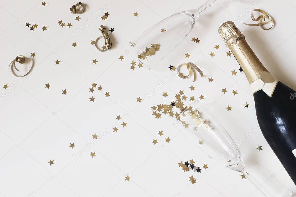 Happy New Year composition. Champagne glasses nad wine bottle with golden confetti stars isolated on white table background. Celebration, party concept. Flat lay, top view. Empty copy space.