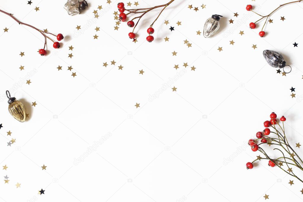 Christmas decorative frame, web banner. Red rose hips, vintage Christmas ornaments and golden confetti stars isolated on white table background. Winter festive decoration. Flat lay, top view.
