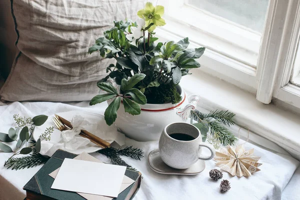 Invierno desayuno bodegón escena. Moody Navidad festiva mesa con cubiertos de oro, taza de café, tarjeta de felicitación, eucalipto y ramas de abeto. Flores de hellebores en macetas en la ventana . —  Fotos de Stock