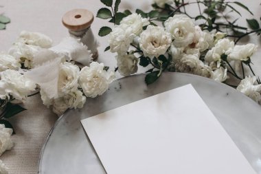 Boş tebrik kartının kapanışı. Huysuz düğün masası modelleme sahnesi. Beyaz gül çiçekleri, plaka ve ipek kurdeleli kadınsı masaüstü kompozisyonu. Seçici odak, bulanık arkaplan.