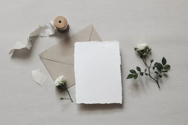 Σύνθεση με στυλ γάμου Μούντι. Γυναικεία επιφάνεια εργασίας mockup σκηνή με λευκά τριαντάφυλλα λουλούδια και φύλλα, μεταξωτή κορδέλα, craft φάκελο και κενό ευχετήρια κάρτα σε γκρι υφή φόντο. Επίπεδο lay, πάνω όψη. — Φωτογραφία Αρχείου