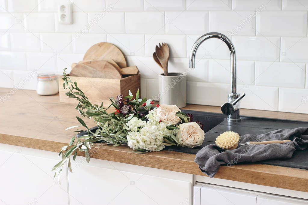 Closeup of kitchen interior. White brick wall, metro tiles, wooden countertops with kitchen utensils. Roses flowers in black sink. Modern scandinavian design. Home staging, cleaning concept.