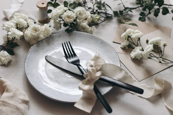 Closeup of festive table setting with black cutlery, white rose flowers, silk ribbons, porcelain dinner plate and envelopes on white table background. Moody wedding party, restaurant eating concept.