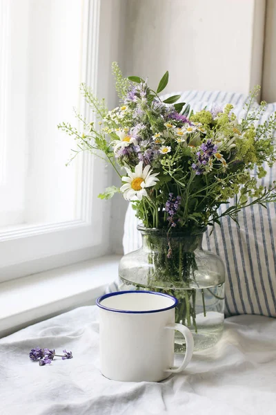 Summer still life scene. Enamel mug, wild flowers bouquet and linen cushion. Vintage feminine photo, rustic design. Floral composition with coffee, lavender, capsella and daisies. Table at window. — Stock Photo, Image