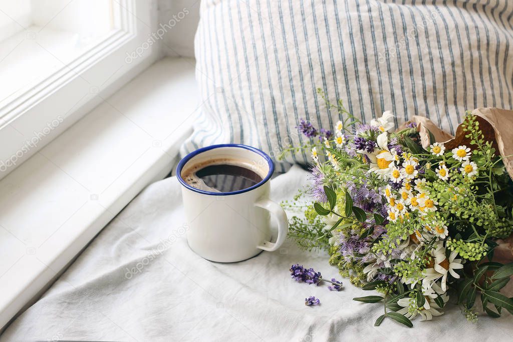 Summer breakfast scene. Enamel mug, wild flowers bouquet and linen cushion. Vintage feminine photo, rustic design. Floral composition with coffee, lavender, capsella and daisies on table at window.