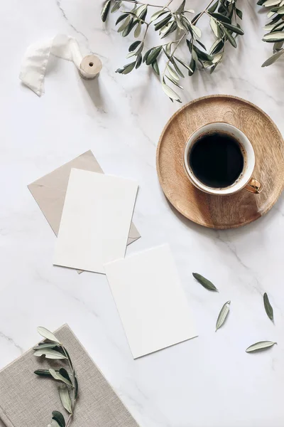 Scène de maquette de papeterie de mariage d'été. Cartes de vœux vierges, assiette en bois, livre, ruban, tasse de café et branches d'olivier. Fond de table blanc avec ombres. Pose verticale à plat féminin, vue de dessus . — Photo