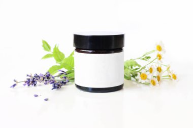 Closeup of dark glass jar with blank paper label. Mint leaves, lavender and feverfew flowers on white table backround. Spa concept. Skin product, moisturizer mockup scene. Cosmetic product packaging. clipart