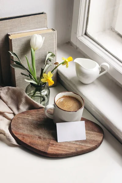 Kellemes húsvét tavaszi csendélet. Névjegykártya mockup jelenet. Egy csésze kávé, könyvek, fa vágódeszka, tejes kancsó és egy váza nárcisz és tulipán virág az ablakpárkányon. Függőleges összetétel. — Stock Fotó