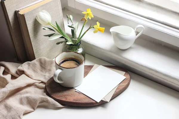 Accogliente Pasqua primavera natura morta. Scheda di auguri scena mockup. Tazza di caffè, libri, tagliere di legno, brocca di latte e vaso di fiori sul davanzale della finestra. Composizione floreale. Giallo narcisi e tulipano . — Foto Stock