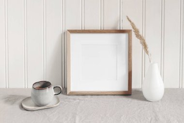 Square empty wooden frame mockup with modern ceramic vase, dry grass, cup of coffee on table. White beadboard wainscot wall paneling background. Scandinavian interior, home design. Art concept.