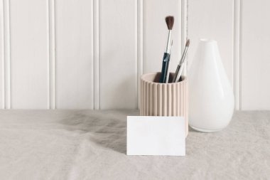 Blank business, place card mockup. Artistic stationery scene with paint brushes, pencils in ceramic holder and glass vase. Creative table composition, white wooden wall background. Front view.