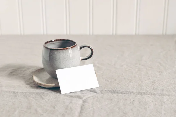 Cup of coffee and blank business, place card. Moody breakfast table stationery mockup scene. Beige linen tablecloth background. Sparse still life composition. Rustic front view, negative space. — Stockfoto