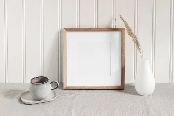 Square empty wooden frame mockup with modern ceramic vase, dry grass, cup of coffee on table. White beadboard wainscot wall paneling background. Scandinavian interior, home design. Art concept. — Stock fotografie