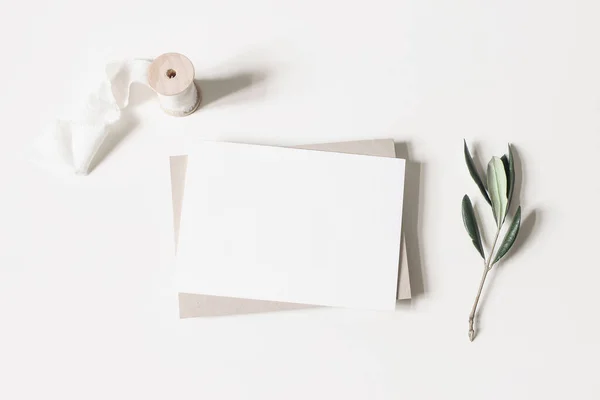 Καλοκαιρινή μακέτα γάμου. Λευκά οριζόντια ευχετήρια κάρτα, φάκελο, κορδέλα και κλαδιά ελιάς που απομονώνονται σε λευκό φόντο τραπεζιού. Θηλυκό μεσογειακό επίπεδο lay, κορυφαία άποψη. — Φωτογραφία Αρχείου