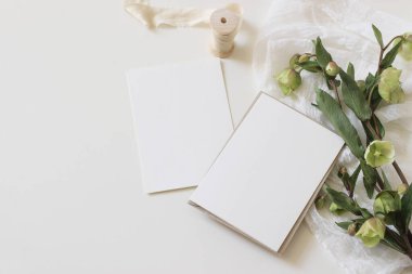 Düğün baharı desenli fotoğraf. Yeşil hellebores çiçekleri, ipek kurdele, muslin bezi ve beyaz masa arkasında boş kağıt tebrik kartları olan kadın masaüstü modelleme sahnesi. Düz yatış, üst görünüm.