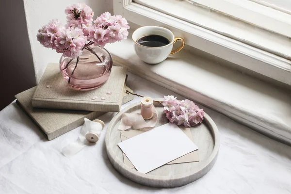 Spring still life scene. Greeting cards mockups, marble tray, cup of coffee, old books. Vintage feminine styled photo. Floral composition with pink sakura, cherry tree blossoms on table near window. — Stock Photo, Image