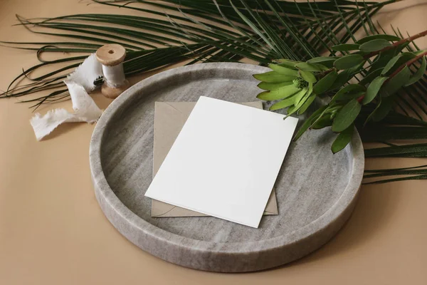 Mock-up scena di nozze tropicali estive di cancelleria. Biglietto d'auguri bianco, busta, nastro e foglie di palma verde e fiori di leucadendro su vassoio di marmo. Sfondo tavolo beige. Vista dall'alto ad angolo alto . — Foto Stock