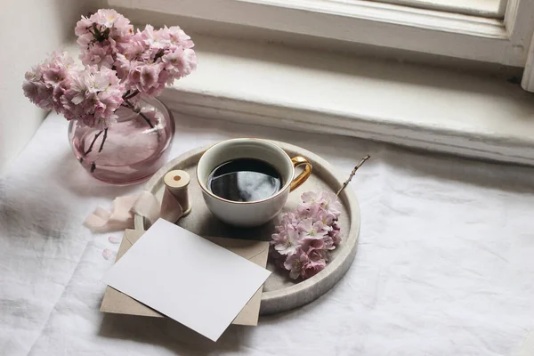 Szene aus dem Frühlingsstillleben. Grußkarten-Attrappen, Umschlag, Marmortablett, Tasse Kaffee. Vintage feminin gestylte Foto. Florale Komposition mit rosa Sakura, Kirschbaumblüten auf dem Tisch neben dem Fenster. — Stockfoto