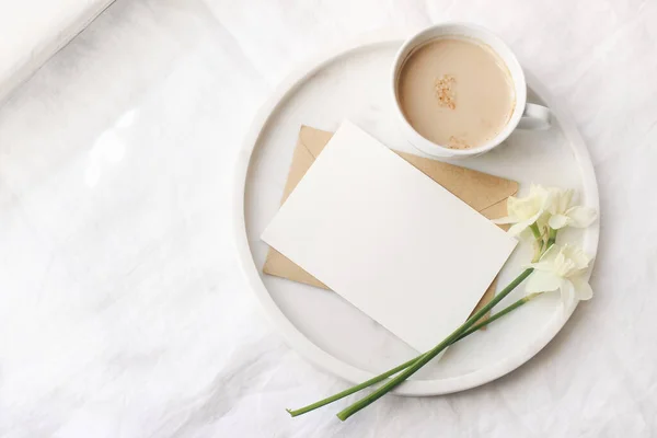 窓の近くのベッドのモックアップシーンで朝食。コーヒー、グリーティングカード、ナルシス、大理石のトレイ上のダフォドールの花のカップ。白いリネンのテーブルクロスの背景。春、イースターの概念。フラットレイアウト、トップビュー. — ストック写真