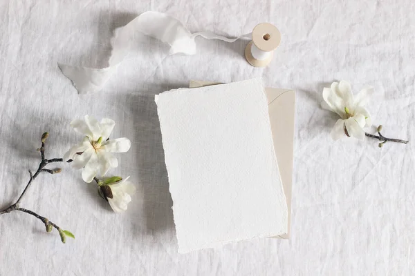 Cena de mock-up de papelaria de casamento. Cartão de saudação vertical em branco, envelope sobre fundo de toalha de mesa de linho com magnólia branca galhos da árvore stellata e fita. Uma vida ainda feminina. Deitado plano, vista superior . — Fotografia de Stock