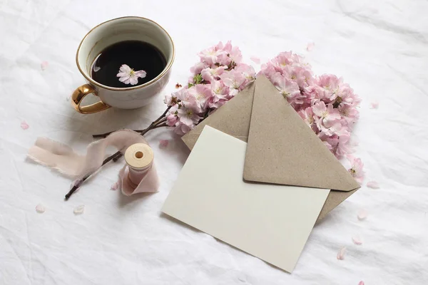 春の静物画。グリーティングカードモックアップ、封筒、白いリネンのテーブルクロスにコーヒーカップ。ヴィンテージのフェミニンなスタイルの写真。ピンクの桜、桜、トップビューと花の組成. — ストック写真