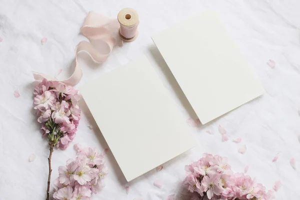 Matrimonio primavera stock foto in stile. Scena femminile di mockup desktop con rami di ciliegio giapponesi in fiore rosa e biglietti di auguri in carta bianca su sfondo bianco della tavola di lino. Posa piatta, vista dall'alto . — Foto Stock