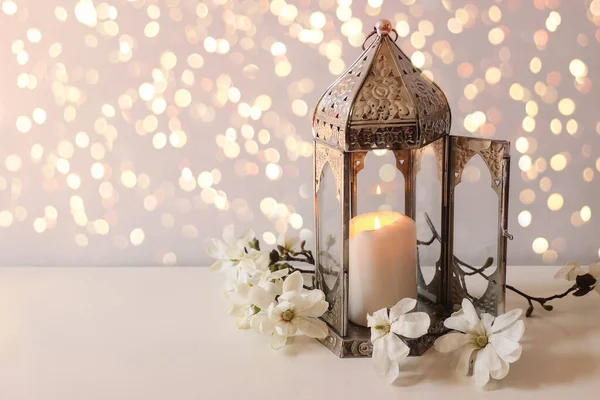 Festive greeting card, invitation for Muslim holiday Ramadan Kareem.Vintage silver Moroccan lantern with glowing candle, white star magnolia branches on table. Glittering bokeh lights. — Stock Photo, Image