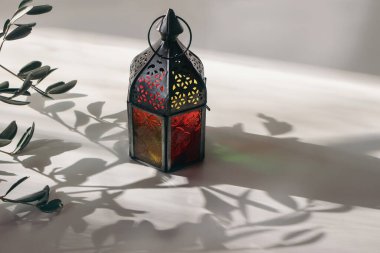 Ramazan Kareem tebrik kartı, davetiye sahnesi. Bulanık zeytin dalları olan renkli Fas feneri. Gündoğumunda beyaz masa, gün batımı ışığı, uzun gölgeler. Kurban Bayramı arkaplanı.