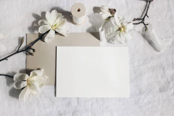 Wedding stationery mock-up scene. Blank horizontal greeting card, envelope on linen tablecloth background. White magnolia stellata tree branches and ribbon in sun. Feminine still life. Flat lay, top