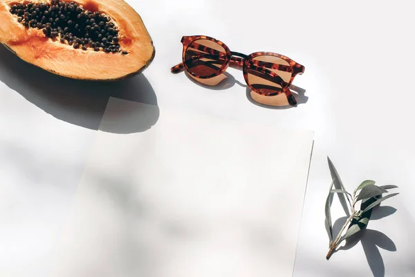 Summer stationery still life scene. Closeup of cut papaya fruit, olive branch and sunglasses in sunlight. White table. Blank paper cards, invitations mockup scenewith harsh shadows. Top view.