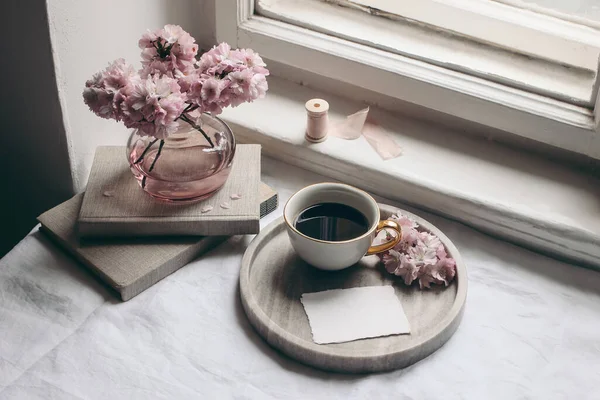 Natura morta primavera composizione. Biglietto d'auguri mockup, vassoio di marmo, tazza di caffè, vecchi libri. Vintage foto in stile femminile. Scena floreale con sakura rosa, fiori di ciliegio sul tavolo vicino alla finestra . — Foto Stock